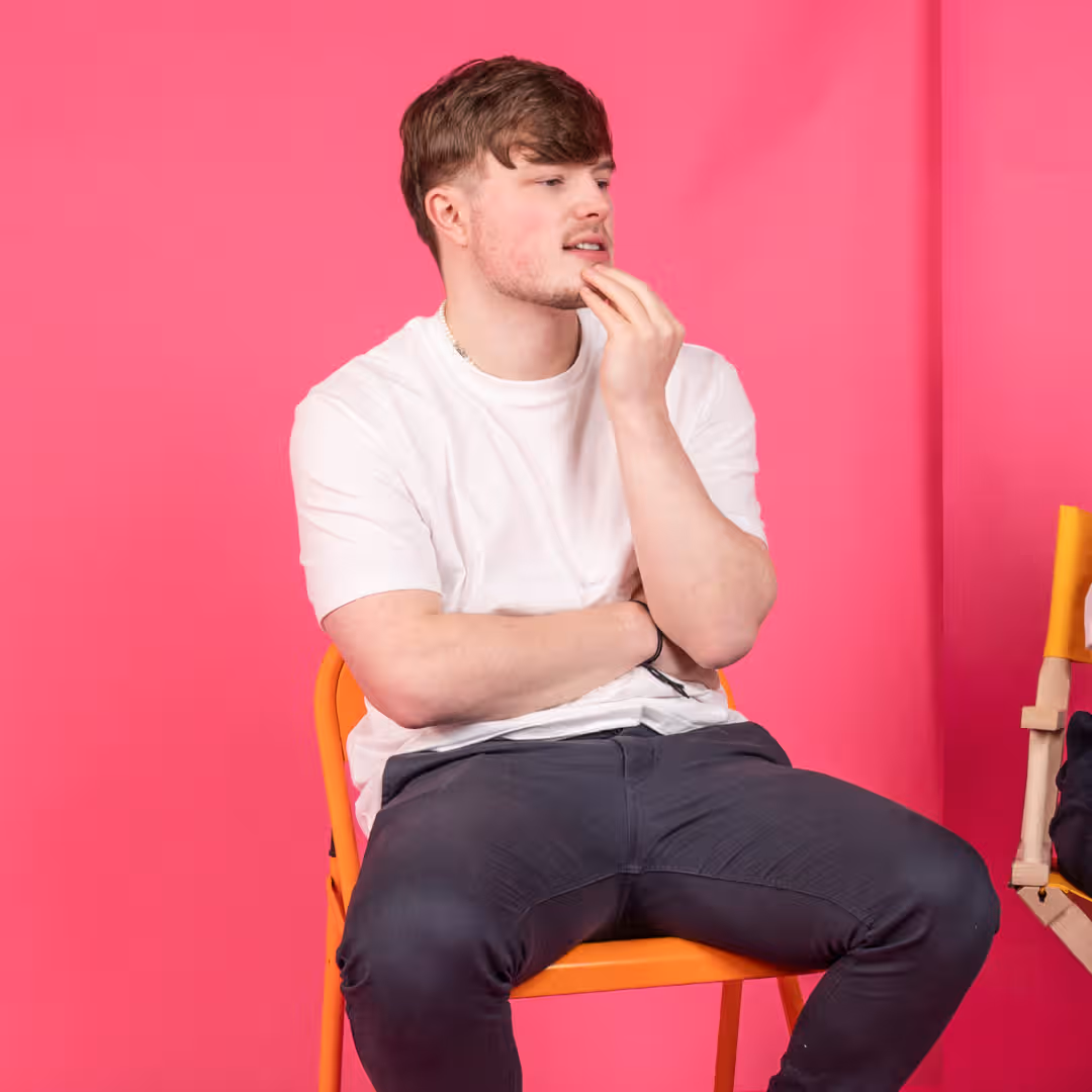 Man in white t-shirt on orange chair, thoughtful pose, vibrant pink studio background