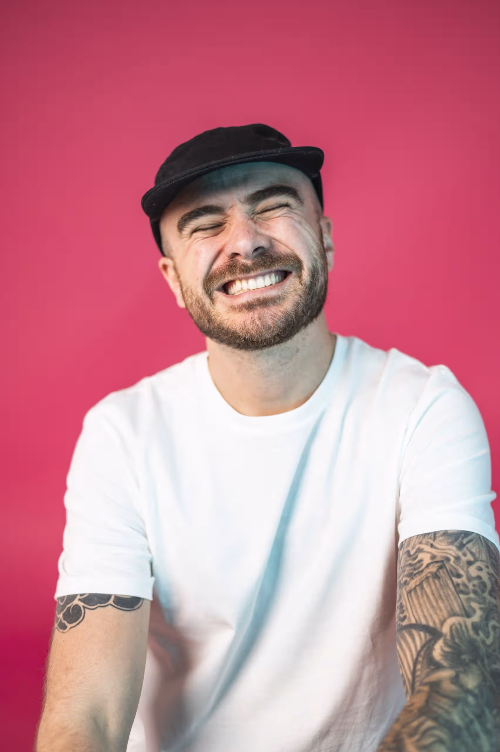 Man smiling broadly, wearing a white t-shirt and black cap, tattooed arm, against a vibrant pink background.