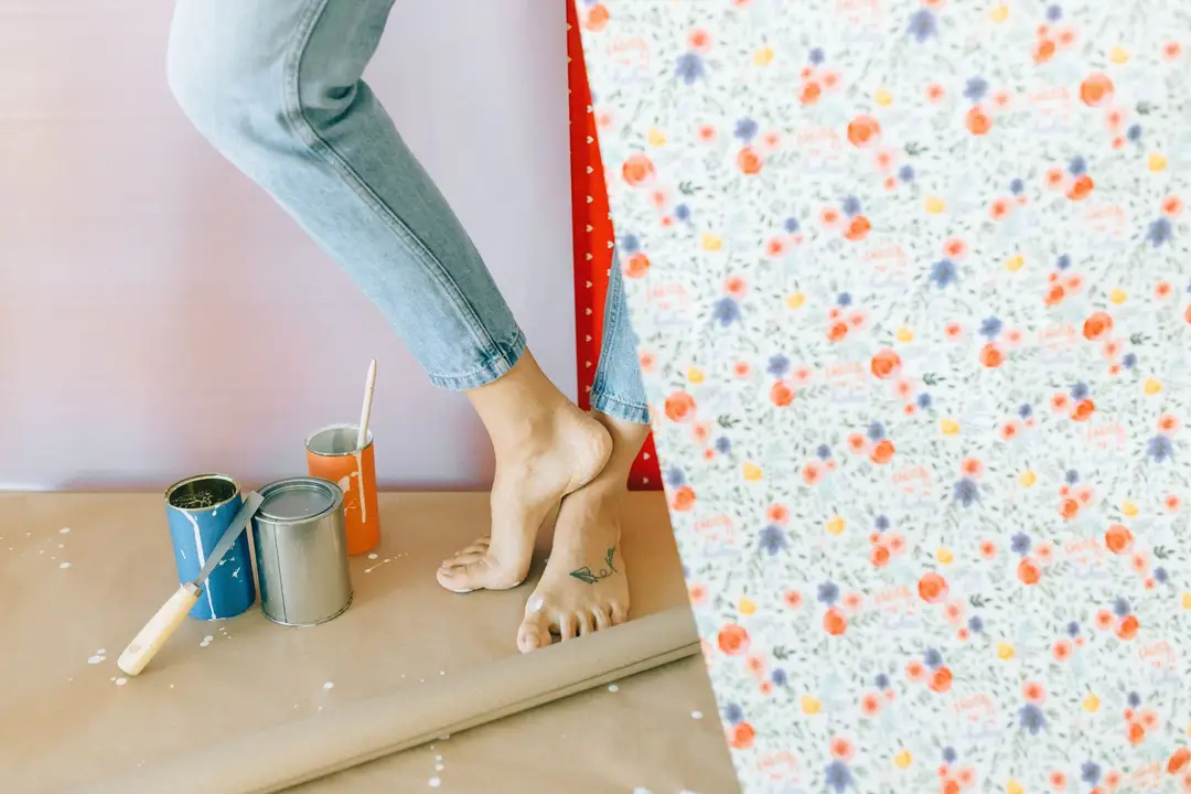 A woman decorates a room with floral wallpaper, surrounded by paint cans and a brush. (Photo: Nataliya Vaitkevich via Pexels)