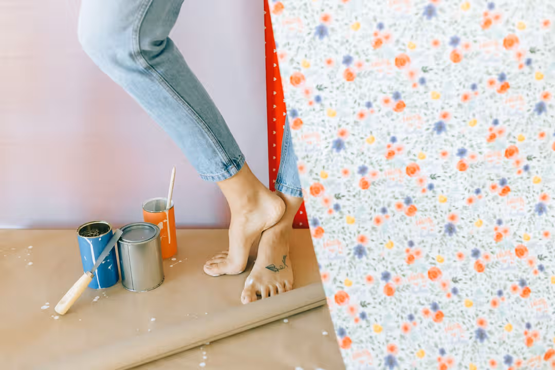 A woman decorates a room with floral wallpaper, surrounded by paint cans and a brush. (Photo: Nataliya Vaitkevich via Pexels)