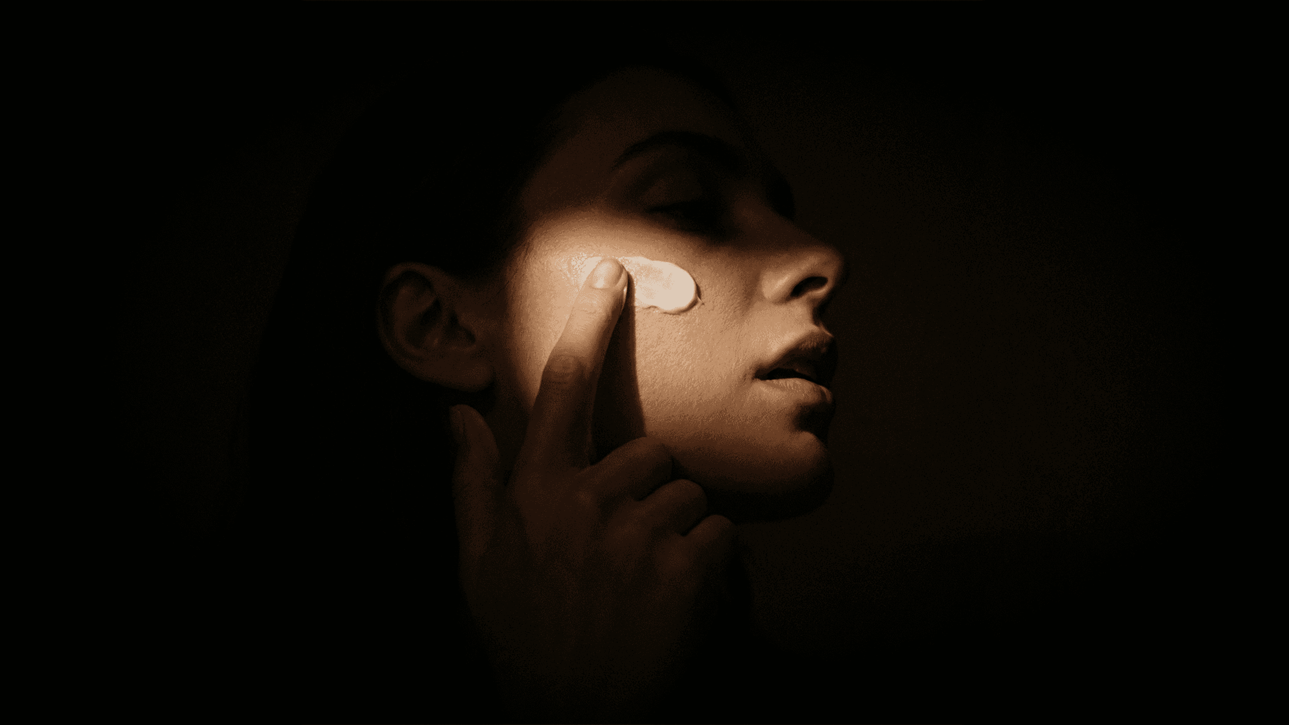 Woman applying skincare cream on cheek in low lighting.