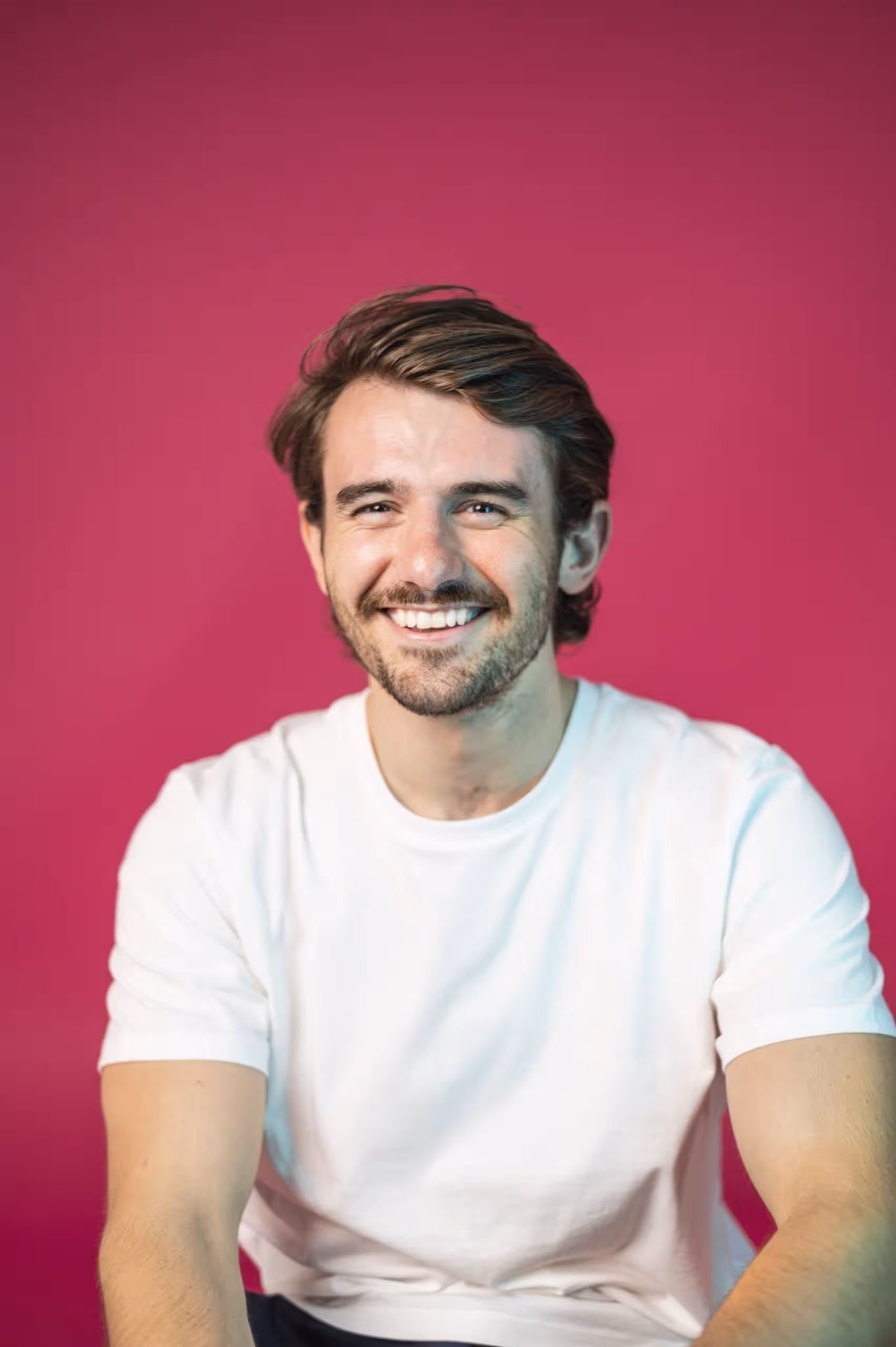 Person smiling, wearing a white t-shirt against a vibrant pink background, representing friendly brand image.