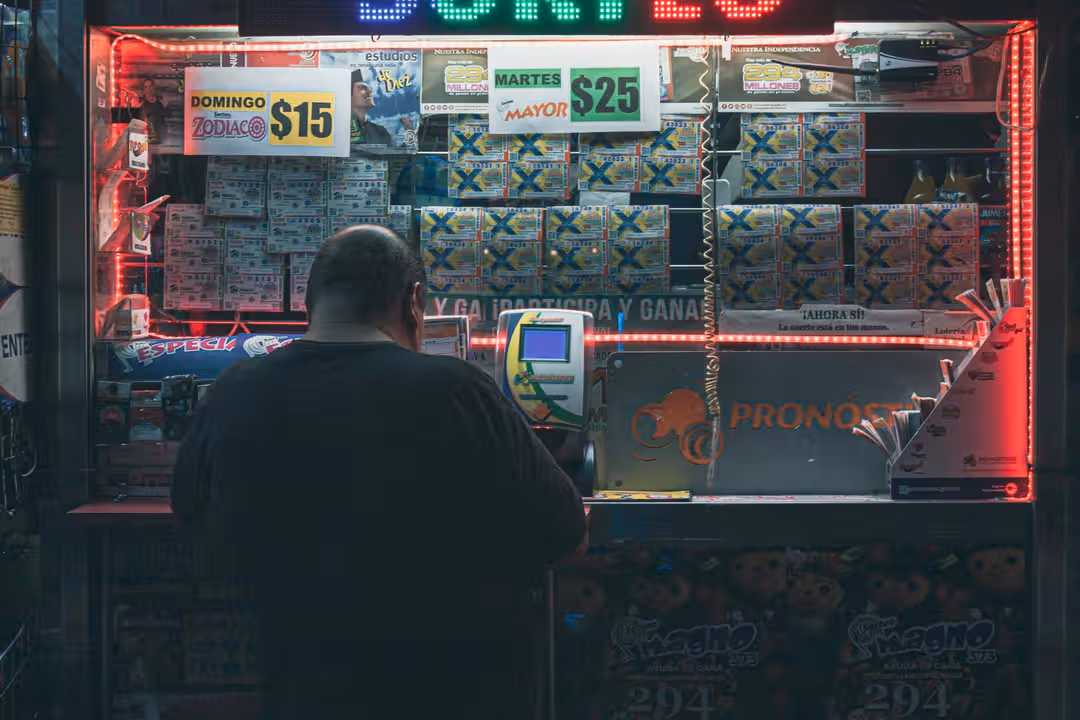 A man purchasing a lottery ticket at an illuminated kiosk in Mexico City, highlighting risk and hope. (Photo: Peaton Hugo via Pexels)