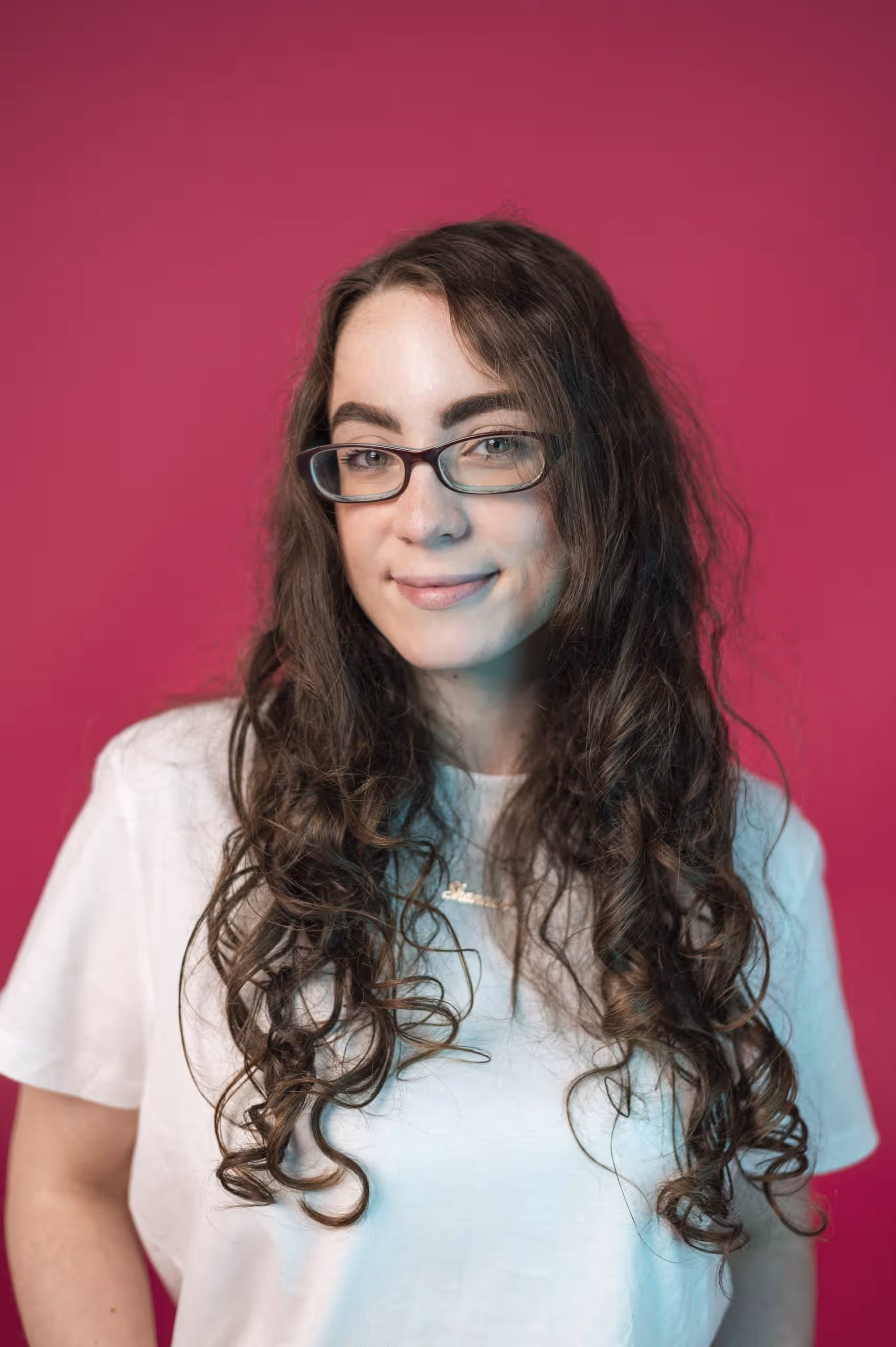 Woman with long curly hair and glasses against a pink background, wearing a white t-shirt.
